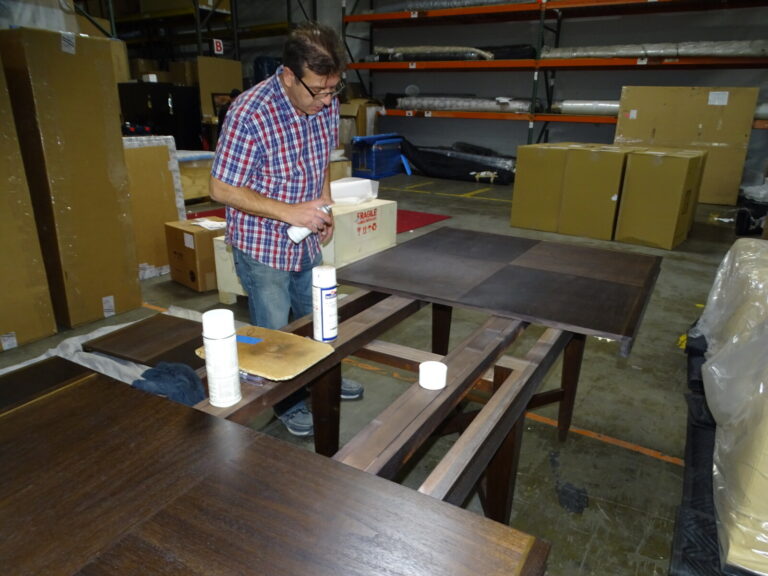 Jeff applying a lacquer topcoat to a baker dining table inside the baker warehouse, demonstrating his hands‑on restoration expertise.