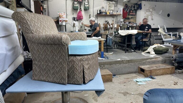 Upholstery workshop with two workers sewing and preparing a partially upholstered armchair among tools, fabrics, and equipment.