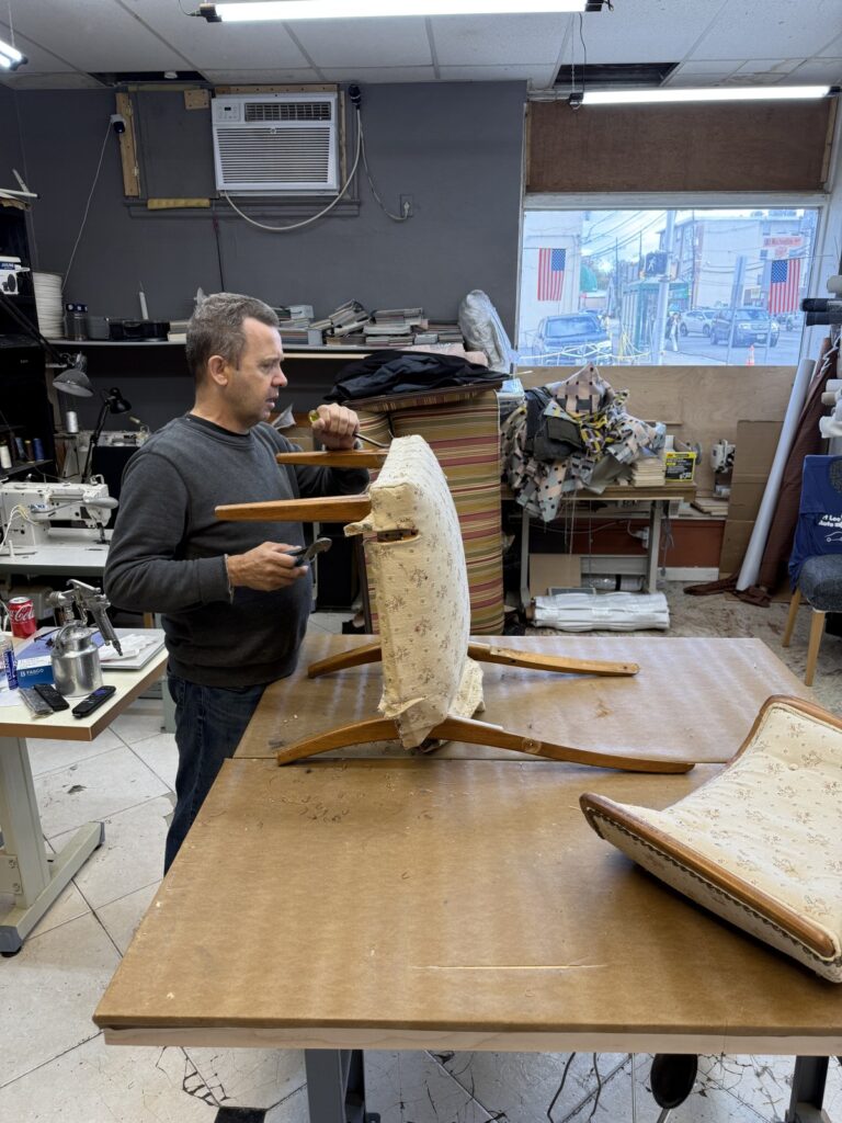 Worker reupholstering a floral-patterned chair on a worktable in a workshop with tools, fabrics, and sewing equipment.