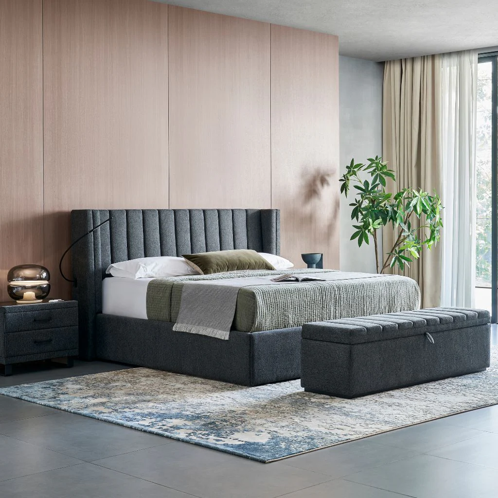 A tall wingback headboard with vertical channel upholstery in a modern bedroom featuring wood wall paneling, a matching upholstered bench, nightstands with lamps, and a large window with sheer curtains.