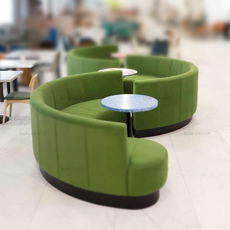 Curved green high‑back upholstered benches arranged around round tables in a modern communal seating area.