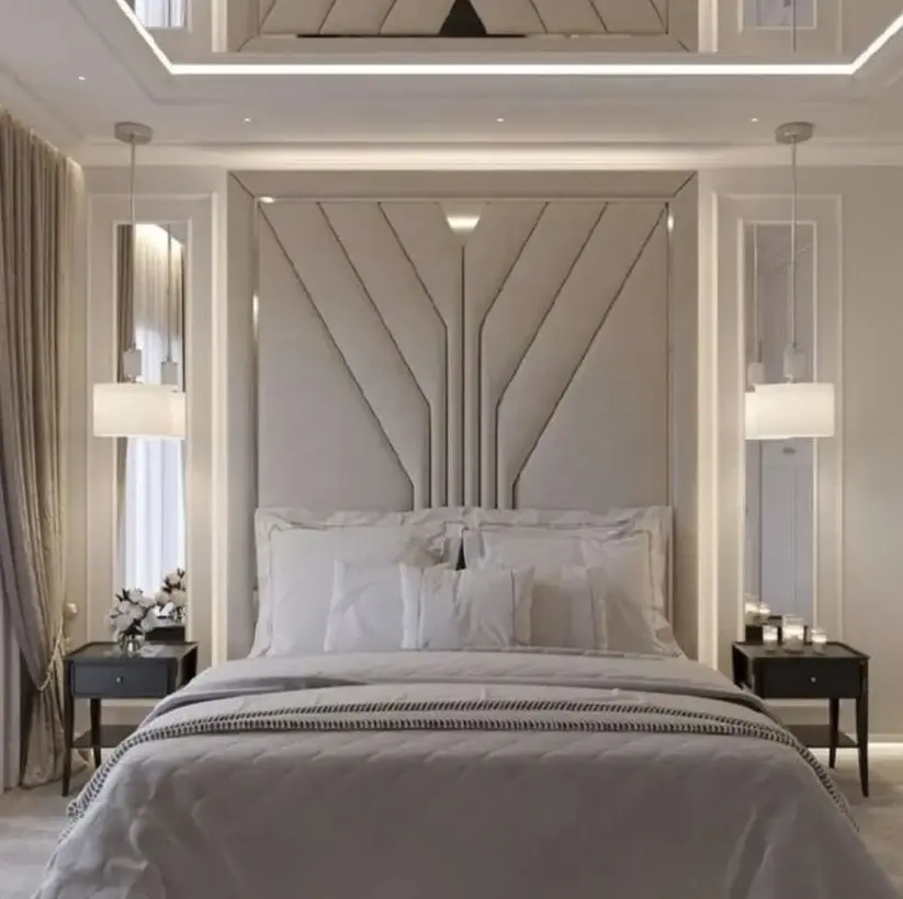 A floor‑to‑ceiling upholstered headboard with soft neutral tones and symmetrical geometric detailing in a modern bedroom with pendant lights, bedside tables, and draped curtains.