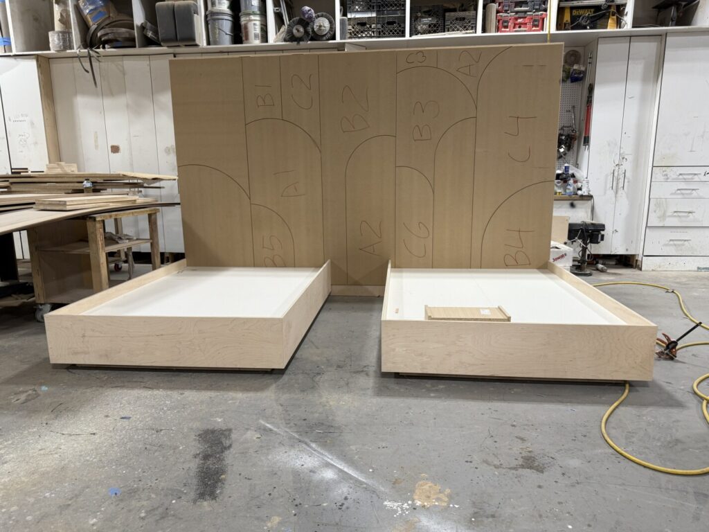 Workshop scene with two bare wood bed frames and large geometric wall panels positioned behind them.