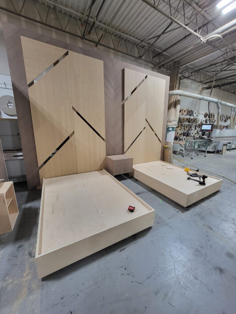 Wood workshop with two wooden bed frames, geometric wall panels, a nightstand, and tools on the floor during assembly.