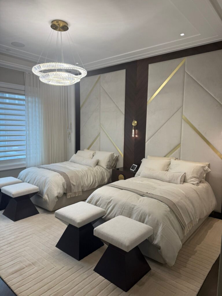 Modern bedroom with two twin beds, geometric wall panels, cushioned stools at the foot of each bed, and a ring‑style ceiling light.