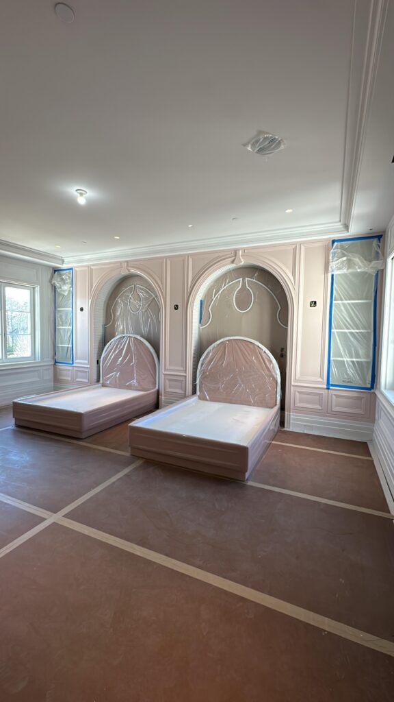 Bedroom under renovation with two wrapped beds, arched wall panels, and protective coverings on floors and built‑ins.