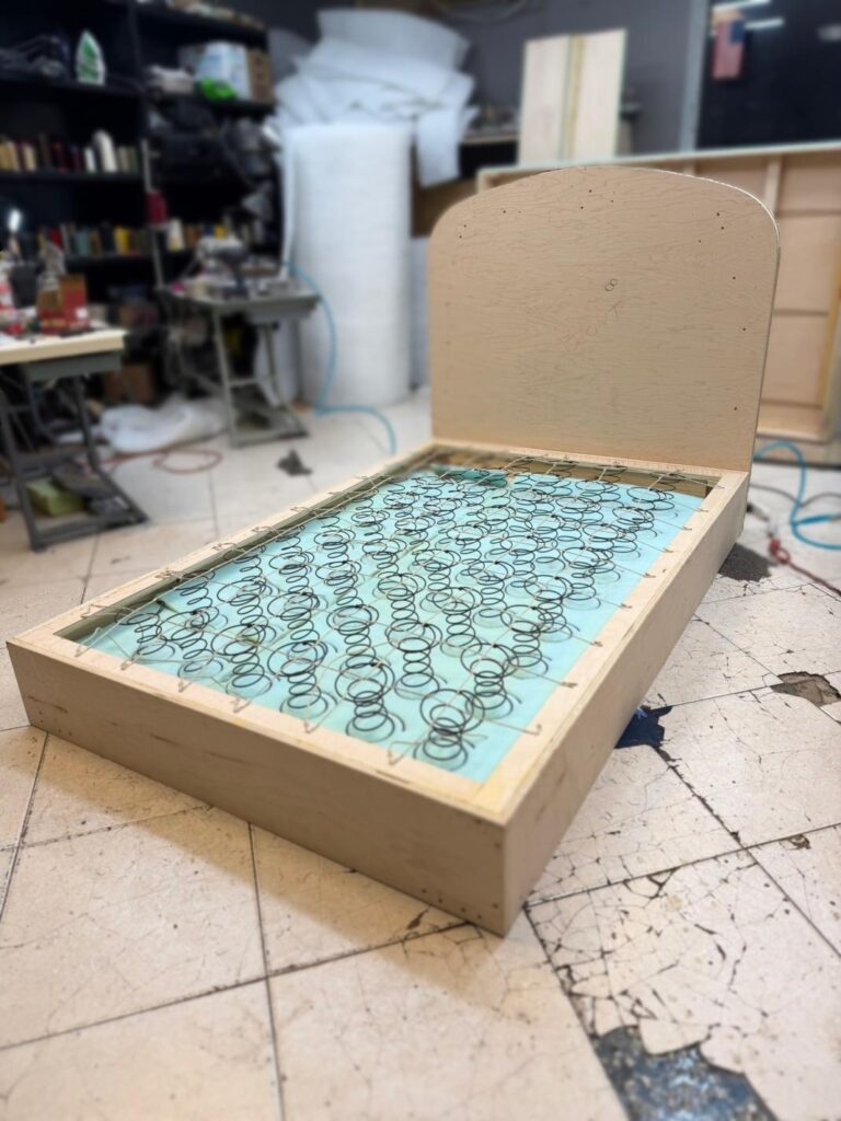 Wooden bed platform with exposed coil springs in a workshop, surrounded by tools, shelves, and upholstery materials.