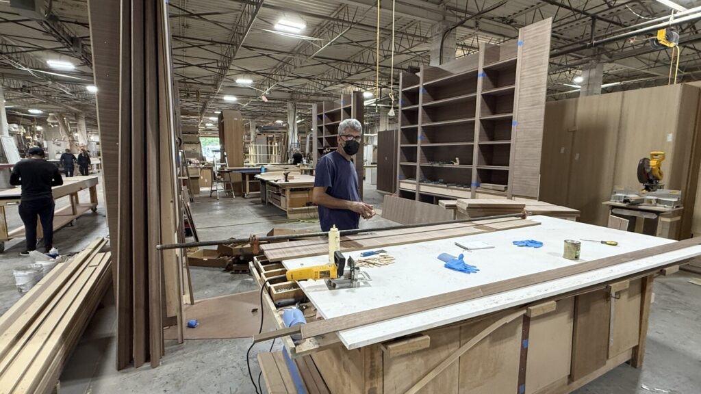 Worker assembling wooden furniture parts at a table in a busy woodworking shop with tools, materials, and equipment in the background.