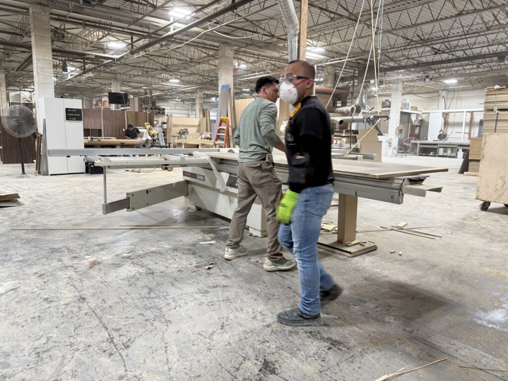 Two craftsmen operating a table saw in the cabinetry shop during the wood‑cutting process.