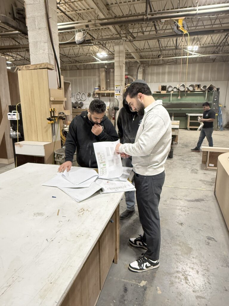 Three people reviewing plans on a worktable in a woodworking shop with tools, machinery, and materials in the background.