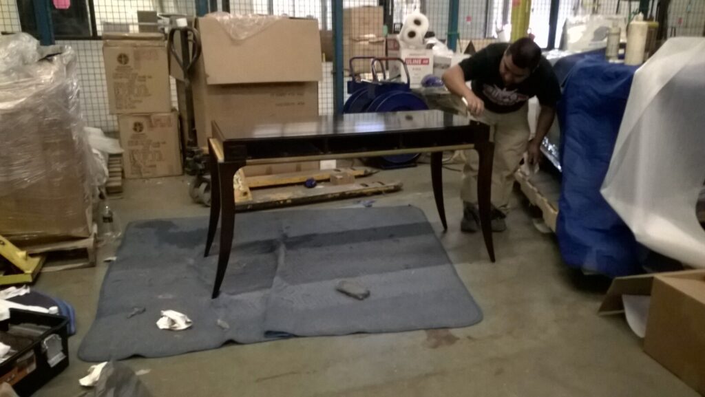 Craftsman inspecting and finishing a dark wooden table in a workshop surrounded by packing materials and equipment.
