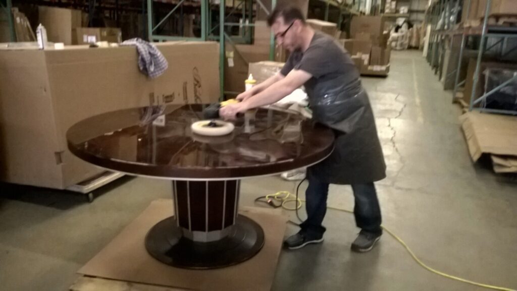 Craftsman machine‑polishing a scratched Baker round dining table in the warehouse to remove surface damage before refinishing.