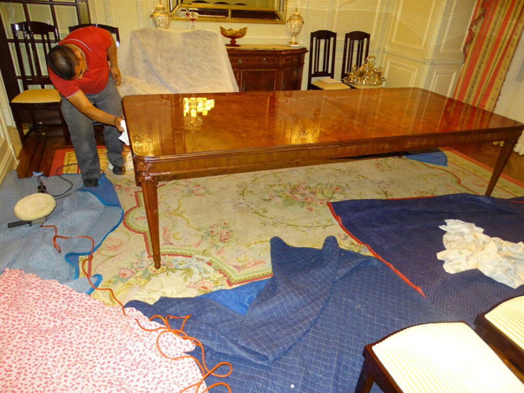 Person polishing the corner of a wooden dining table in a furnished room with protective blankets on the floor