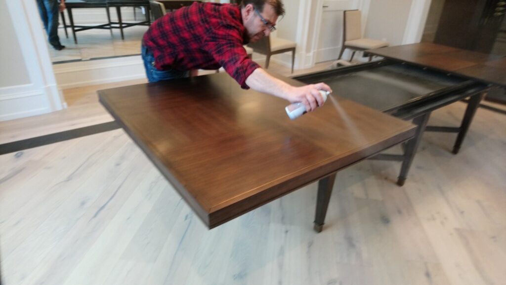 Craftsman spraying a fresh top coat on a Barbara Barry for Baker Larchmont Extension Table during on‑site refinishing in the client’s home.