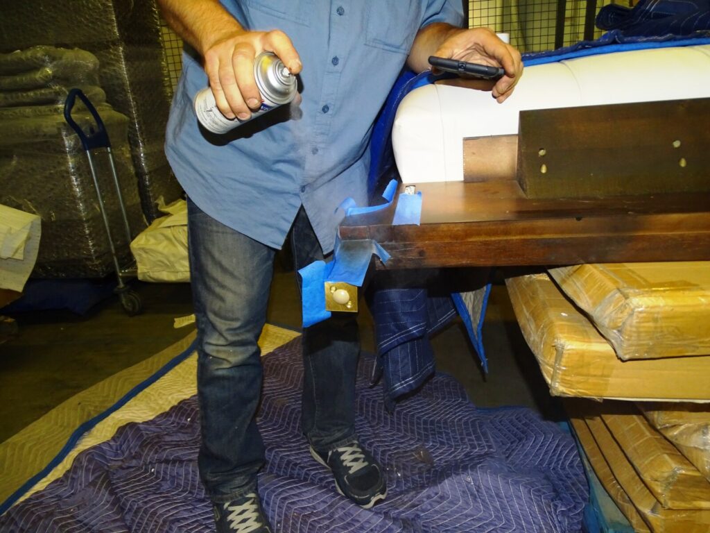Person spraying a masked furniture surface with a can while the piece rests on a padded blanket in a warehouse setting.