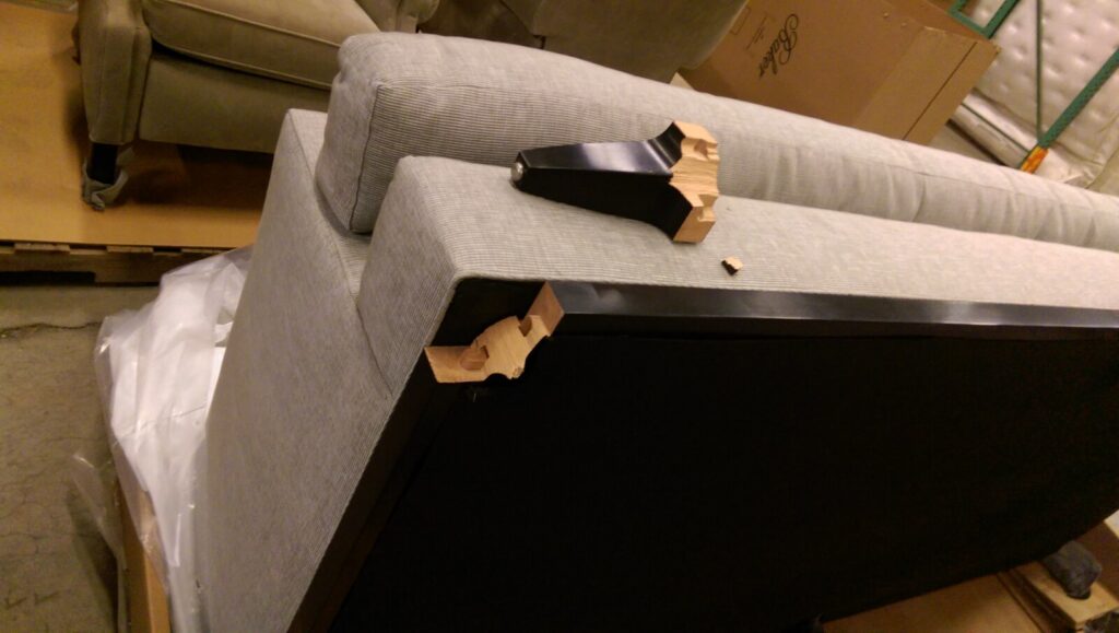 Underside of a sofa with a detached wooden leg resting on the frame and a broken wood piece near the mounting slots