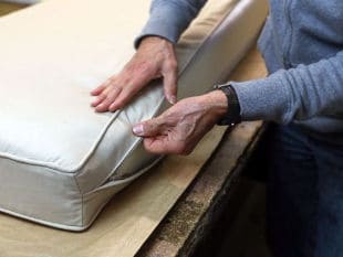 Closeup view of a person’s hands working on the edge of a lightcolored cushion, adjusting the zipper and seam while the cushion rests on a wooden table.
