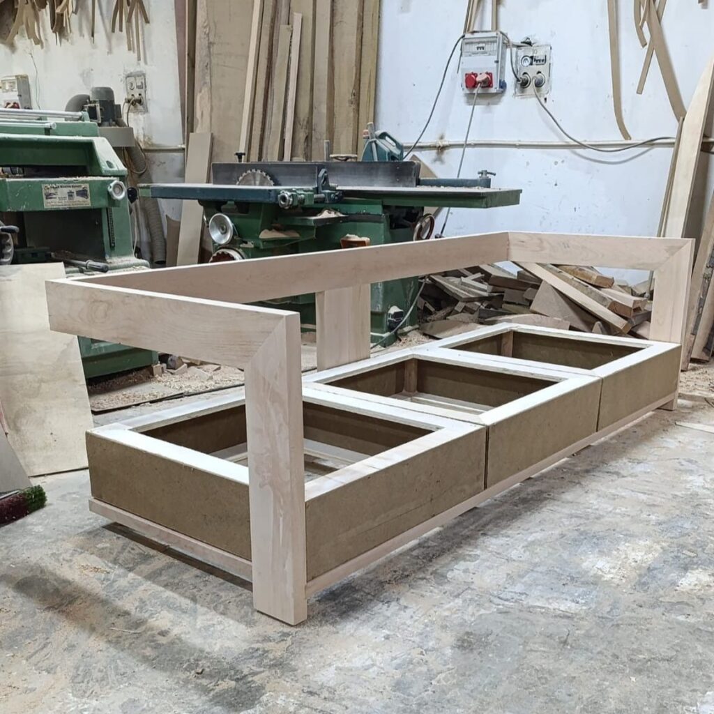 Bare wood sofa frame under construction in a woodworking shop, showing seating support and armrest structure.