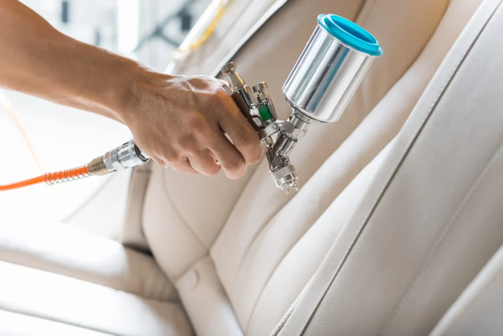 Technician using a spray gun to clean or restore a light‑colored leather car seat.