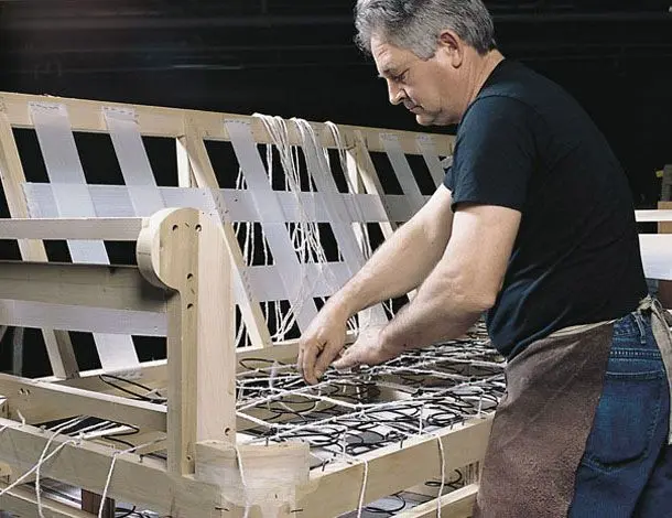 Person working on a wooden furniture frame, securing crisscross webbing and springs while wearing a dark shirt and apron.