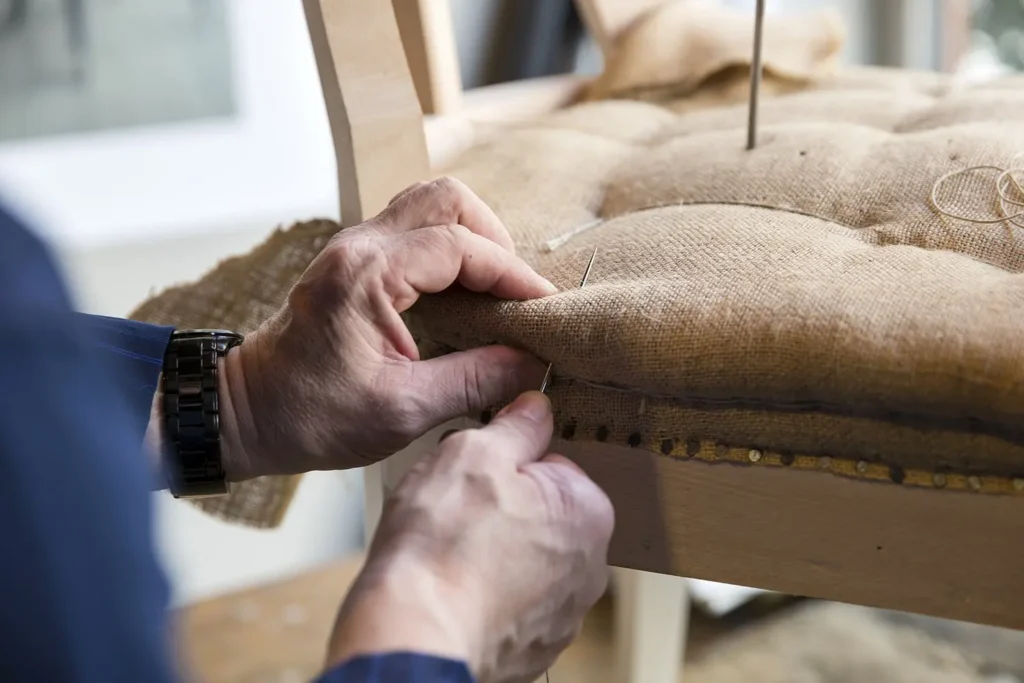 Craftsman hand‑stitching the seat top over coil springs on an antique chair during detailed upholstery restoration.