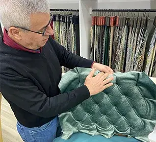 “person handling tufted fabric in a workshop with rolls of material in the background.