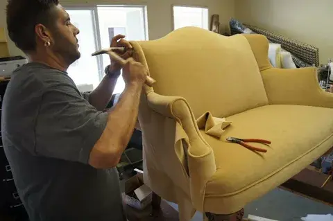 Craftsman reupholstering a beige sofa, securing fabric along the armrest with tools visible on the seat in a bright workshop