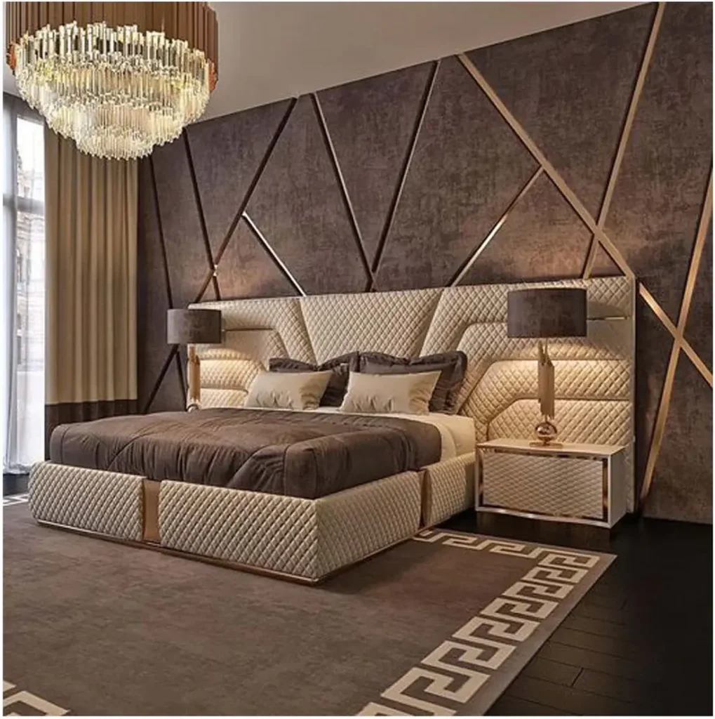 A wall‑to‑wall headboard with quilted upholstery and diagonal gold geometric accents in a luxury bedroom featuring a chandelier, bedside tables with lamps, and a patterned rug.