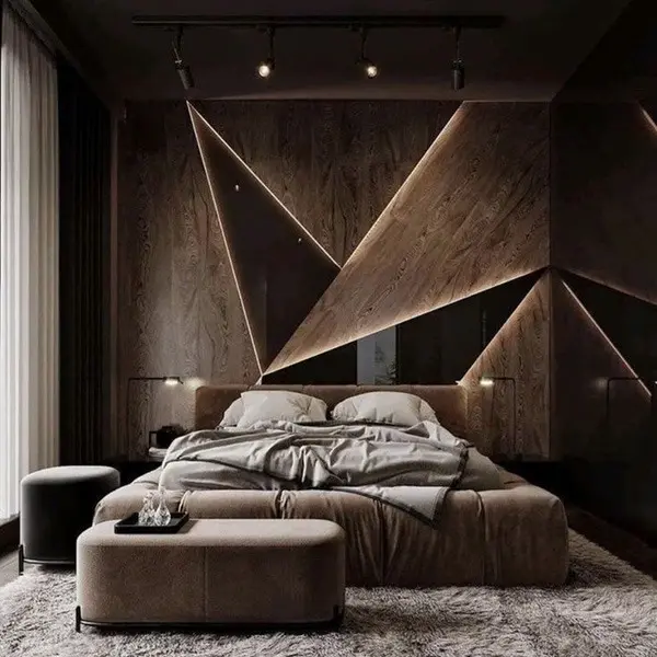 A modern wall‑mounted headboard made of geometric wooden panels with integrated lighting behind a neutral upholstered bed, featuring two ottomans, a textured rug, and soft ambient bedroom lighting.