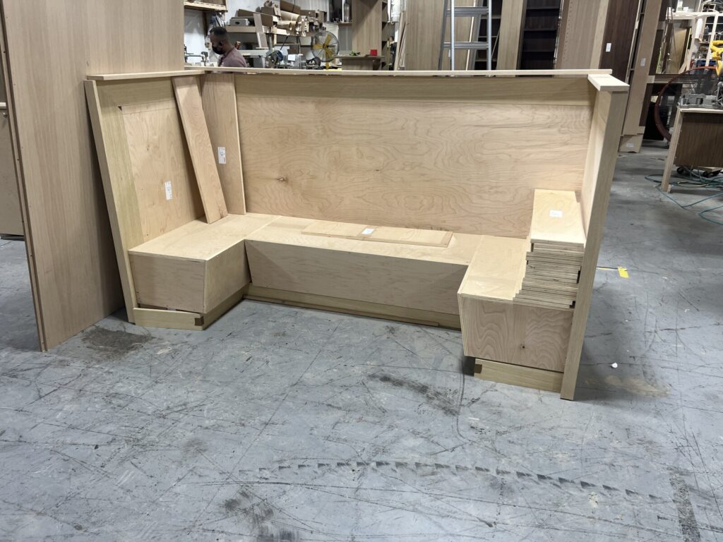 U‑shaped plywood banquette frame in a workshop, with a high backrest and additional panels stacked nearby.