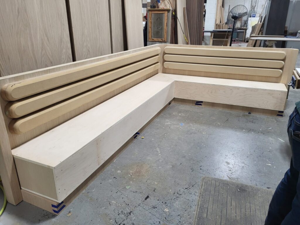 Custom L‑shaped wooden banquette frame with rounded slat backrest in a workshop during fabrication.