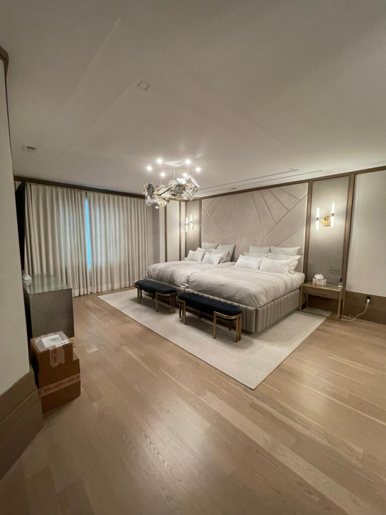 Modern bedroom with two side‑by‑side beds, white bedding, black benches with gold accents, geometric upholstered wall panels and the headboard, chandelier, and floor‑to‑ceiling curtains