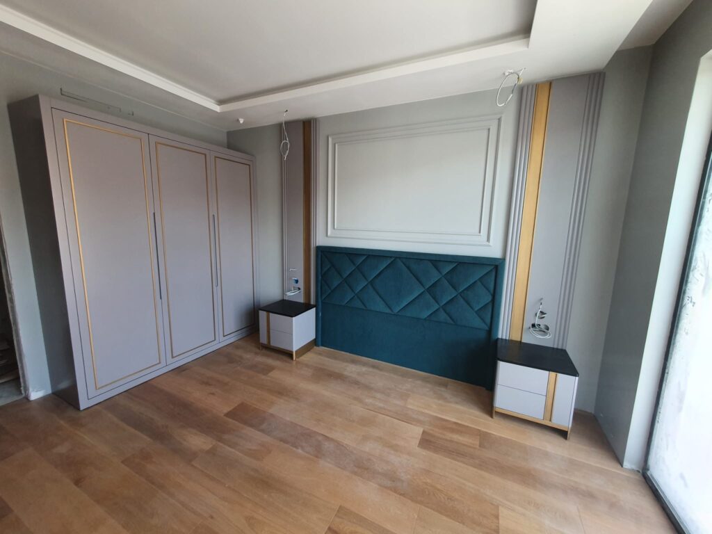 Bedroom under construction with a teal diamond‑tufted upholstered headboard installed between two nightstands, built‑in wardrobe, and paneled wall.