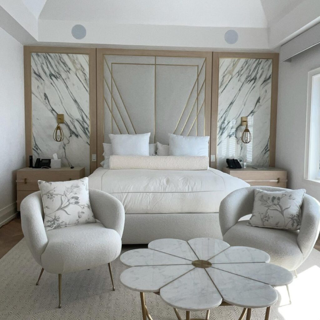 Luxury bedroom with upholstered brass-inlay wall panel, marble wall panels, bouclé chairs, and a marble cocktail table.