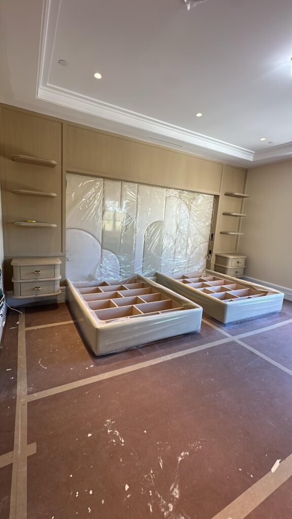 Bedroom under construction with two upholstered bed frames, padded headboard wrapped in plastic, and built‑in shelves on both sides.