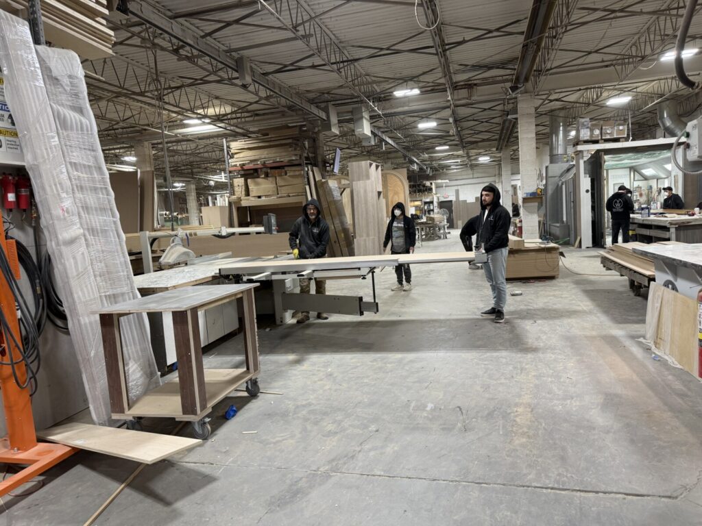 Workers handling large wood panels in a spacious woodworking shop with tools, machinery, and stacked materials visible throughout the space.