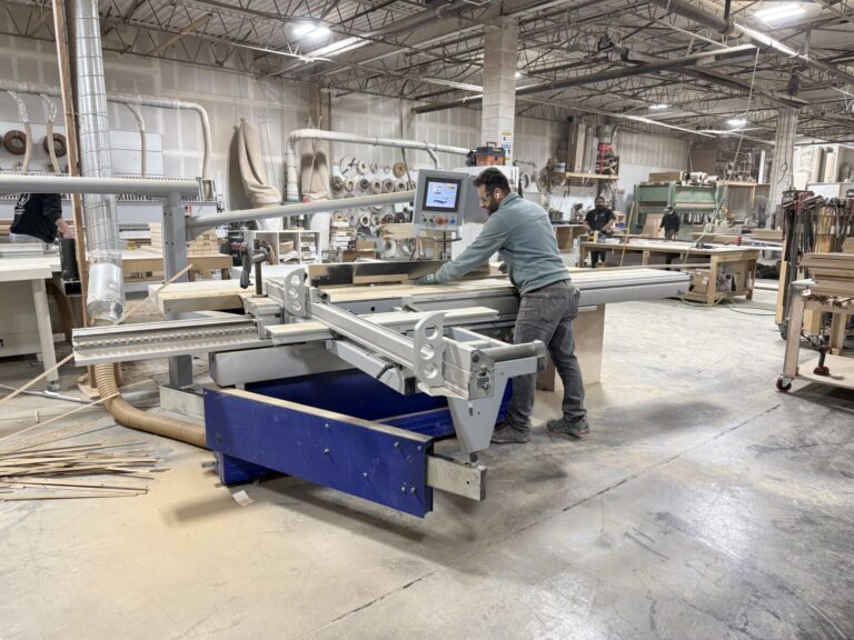 Worker using a sliding table saw in a woodworking shop with wood panels, tools, and industrial lighting.