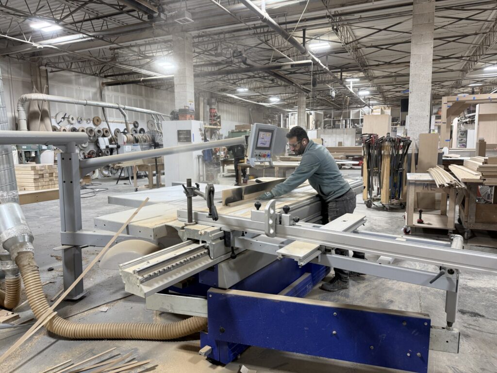 Worker operating a sliding table saw in a woodworking shop, with wood panels, tools, and sanding wheels visible in the background.