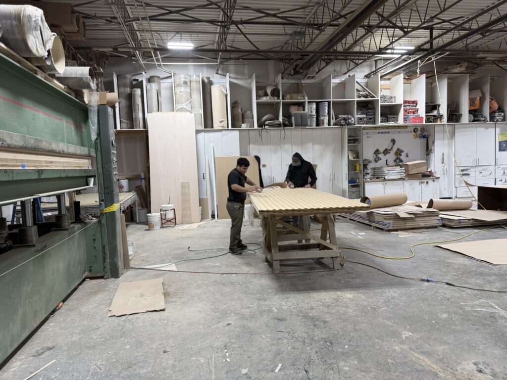 Two workers assembling cabinetry on a shop table with plywood and tools visible in the workshop.