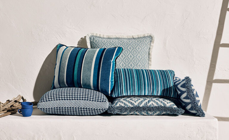 Assorted blue patterned throw pillows arranged on a white bench with driftwood and a small blue cup.