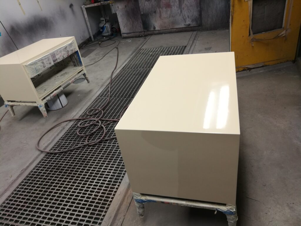 Custom white‑lacquer Barbara Barry bedside table in the spray room with the lacquer finish being built to a smooth, even surface.