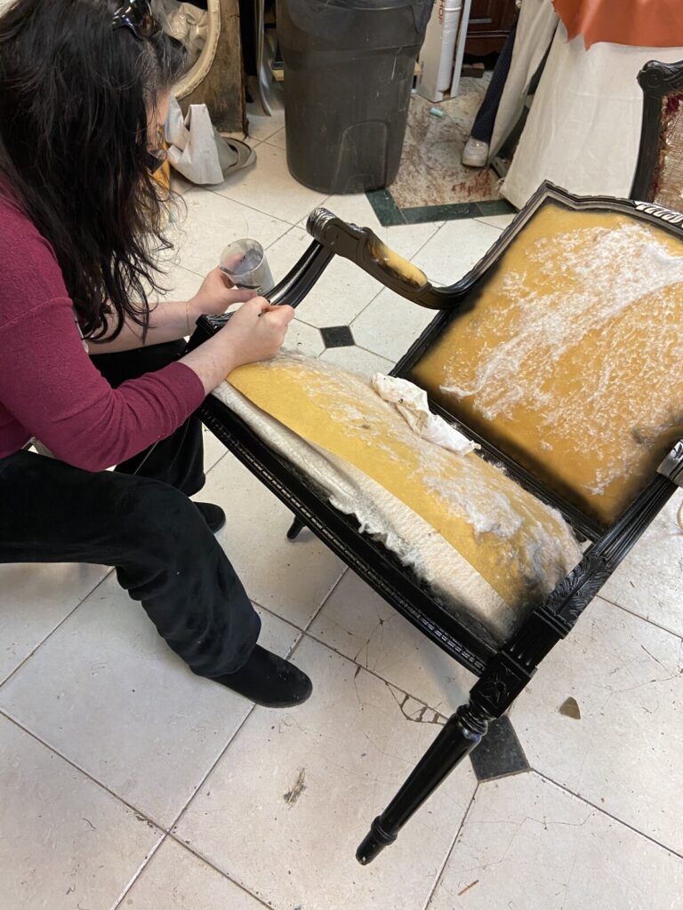 Antique black chair frame in the workshop with a craftsman detailing the silver leaf during restoration.