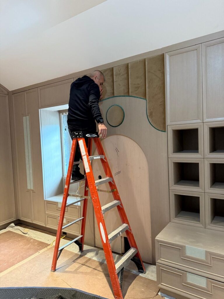 Installer mounting a custom upholstered wall panel while standing on a ladder in a client’s home.