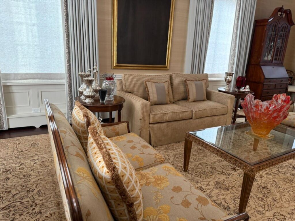 Traditional living room with a beige upholstered sofa, floral settee, glass‑top coffee table, and patterned area rug.