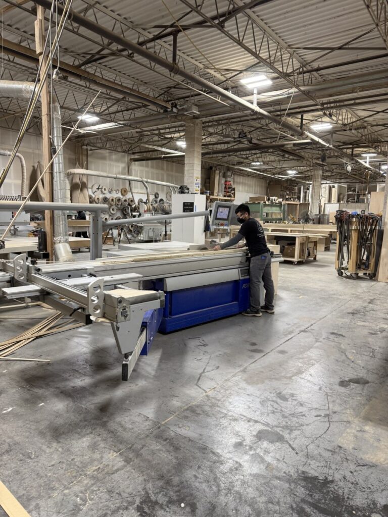 Person operating a large blue CNC woodworking machine in a workshop with wood panels, workbenches, and industrial lighting