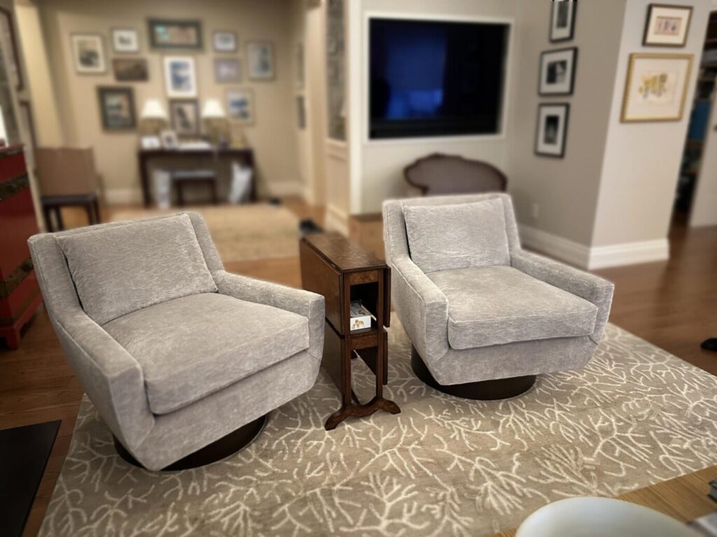 Two light gray swivel armchairs on a patterned rug with a narrow wooden side table between them, in a living room with a wall-mounted TV, framed photos, and a hallway in the background.
