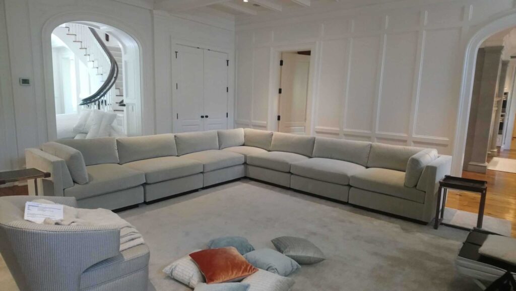 Large light gray L‑shaped sectional sofa on a light carpet in a living room with white paneled walls, arched doorways, a staircase in the background, and a striped armchair with a blanket and pillows on the floor nearby.