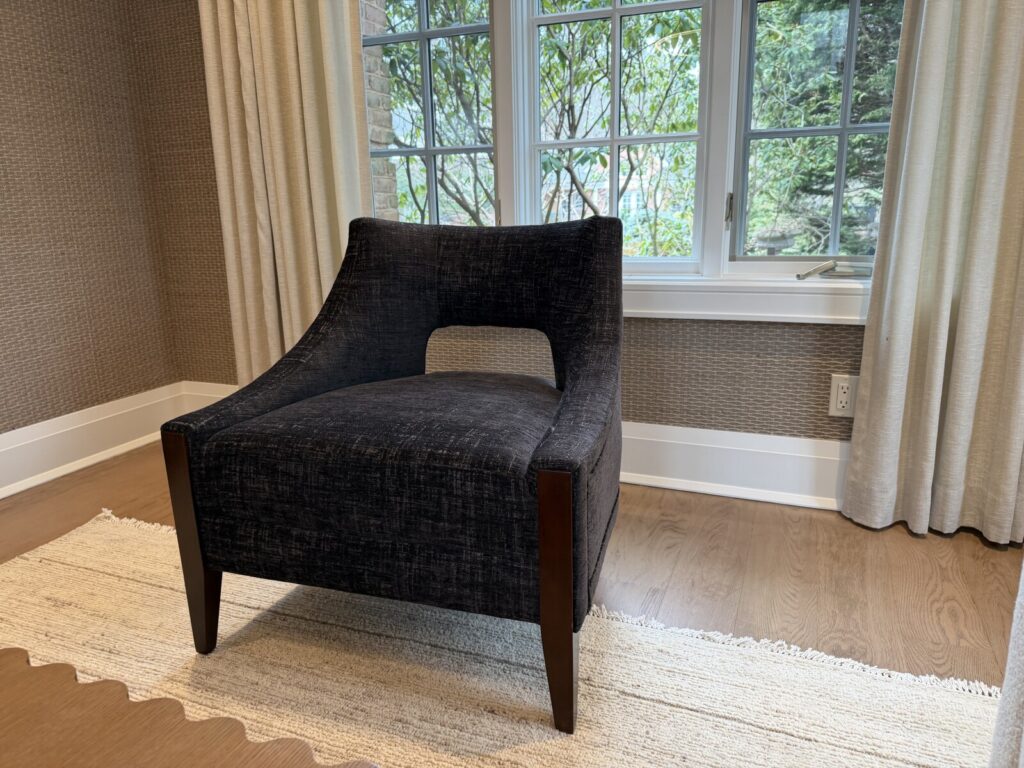 Dark upholstered lounge chair with angled wooden legs positioned on a light wood floor near a window with white curtains.