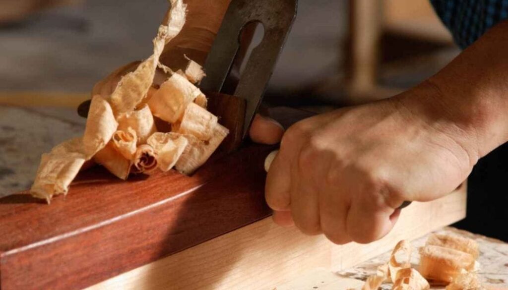 Person using a hand plane to shave curled wood shavings from a dark wooden board.