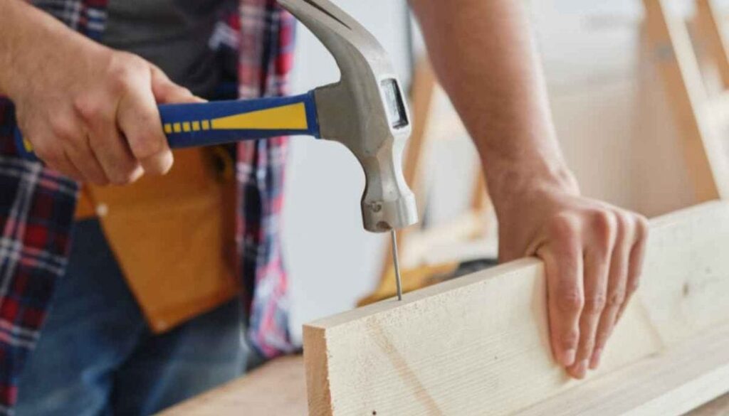 Person hammering a nail into a light wooden board while holding the board steady with the other hand.